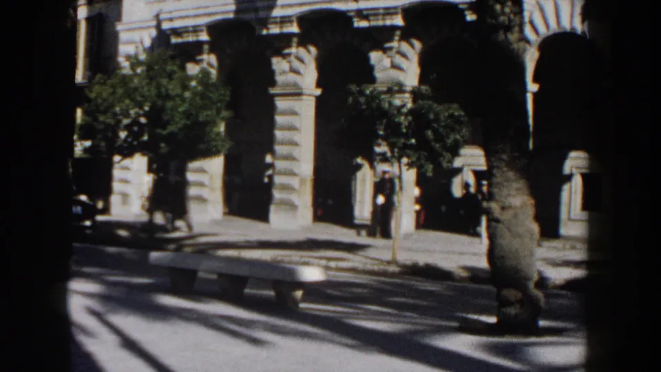 Late-frame view of a 1960 Italy street with an arched beige stone building, pedestrians, trees, bench, and sunlit roadway.