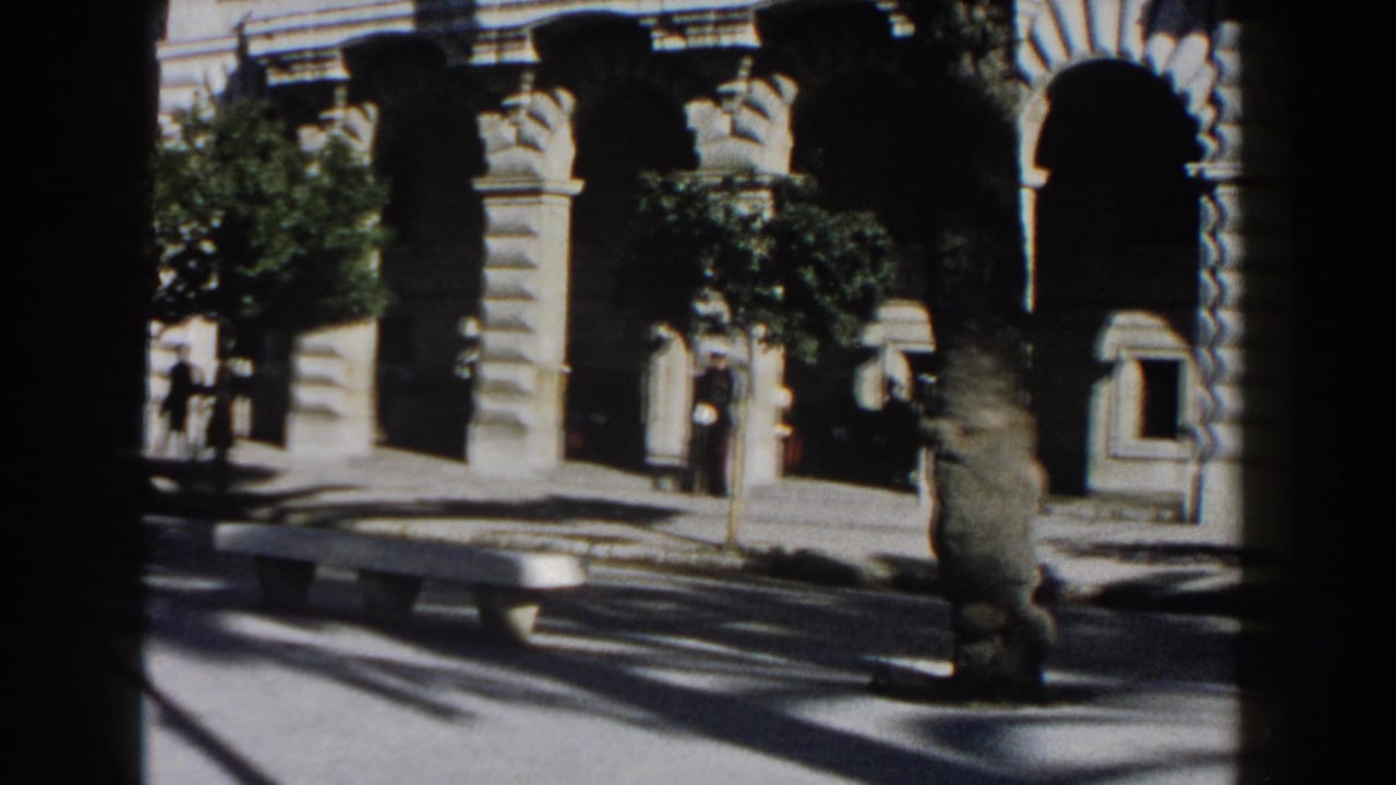 1960 Italy 8mm view of a stone arcade or church entrance with palm trees, shadows, and distant pedestrians.