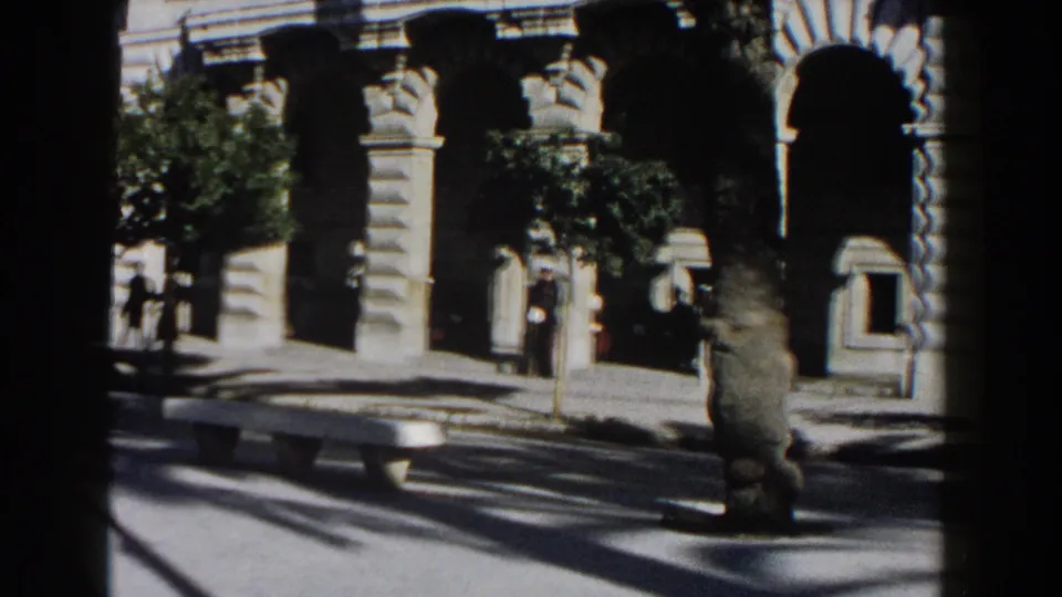 1960 Italy 8mm view of a stone arcade or church entrance with palm trees, shadows, and distant pedestrians.