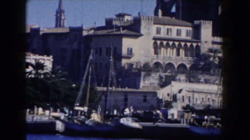 1960 Italy 8mm view of waterfront buildings with arches, a church spire, palm trees, water, and small boats.