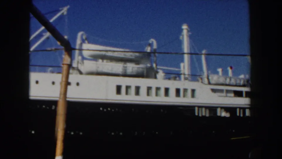 1960 Italy 8mm frame showing the side of a docked passenger ship with rows of windows, deck fittings, and foreground railings.