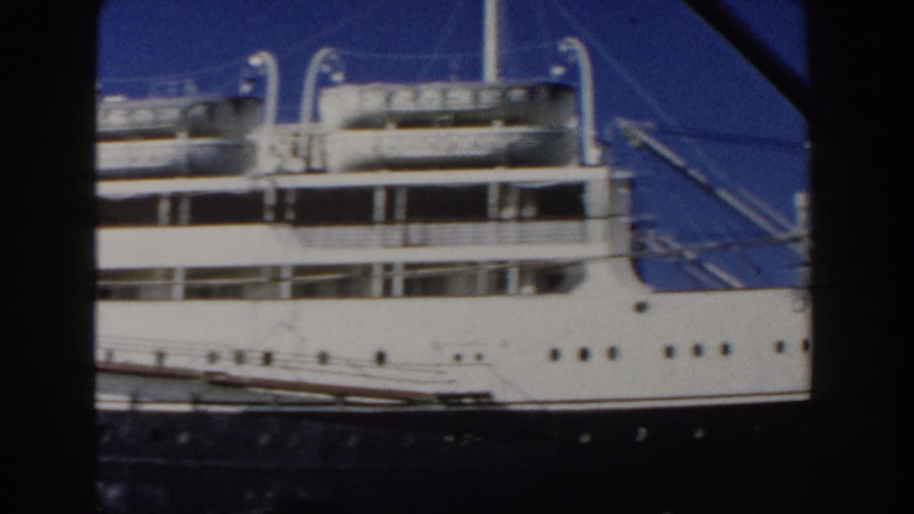 Side of a docked passenger ship with lifeboats, windows, and dark hull under blue sky.