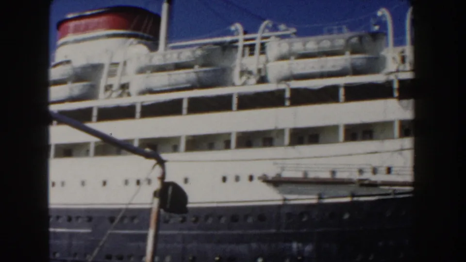 1960 Italy 8mm frame of a docked passenger ship midsection, with red-topped funnel, lifeboats, railings, and rigging lines crossing the view.