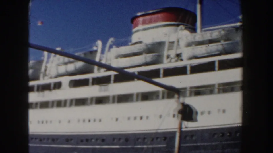 1960 Italy 8mm frame showing a closer port-side view of the ship's upper decks, funnel, lifeboats, windows, and diagonal dockside line.