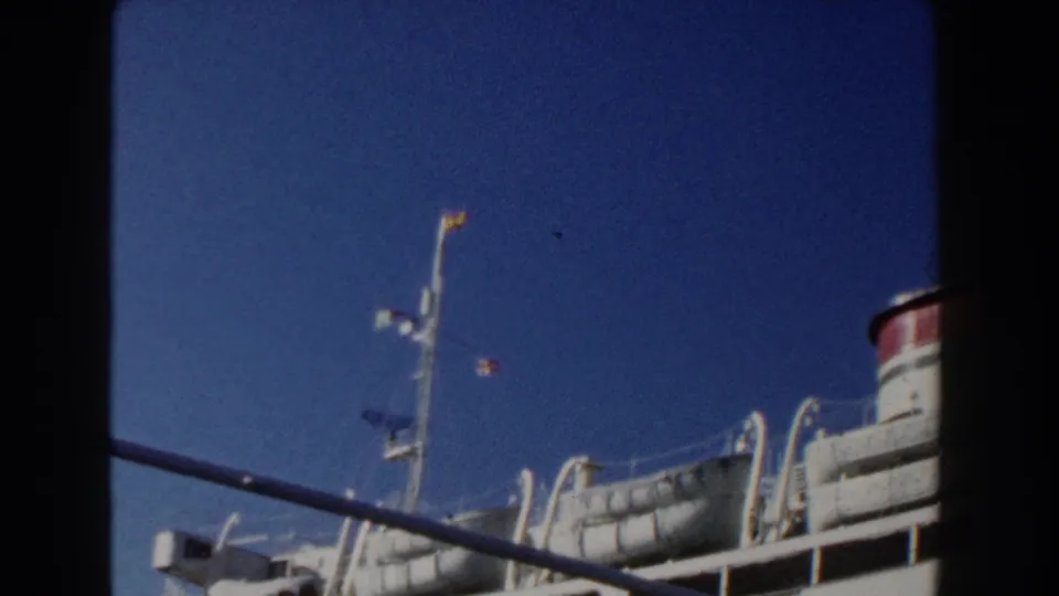 1960 Italy 8mm frame showing a white ocean liner with upper decks and lifeboats viewed from a nearby vessel.