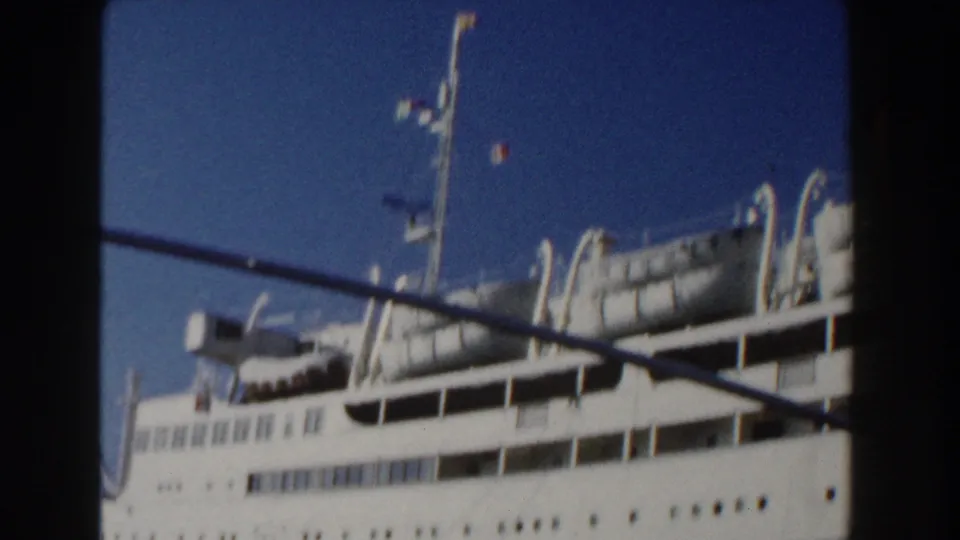 White passenger liner in bright sun, with lifeboats and a tall mast visible against the blue sky.