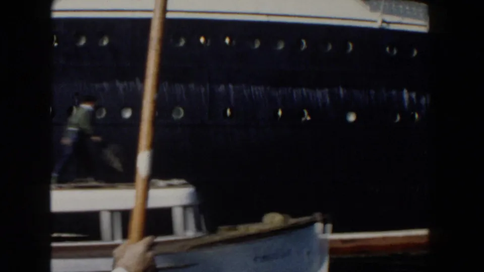 Home-movie view of the liner's dark hull above a small vessel, with one distant person visible on deck.