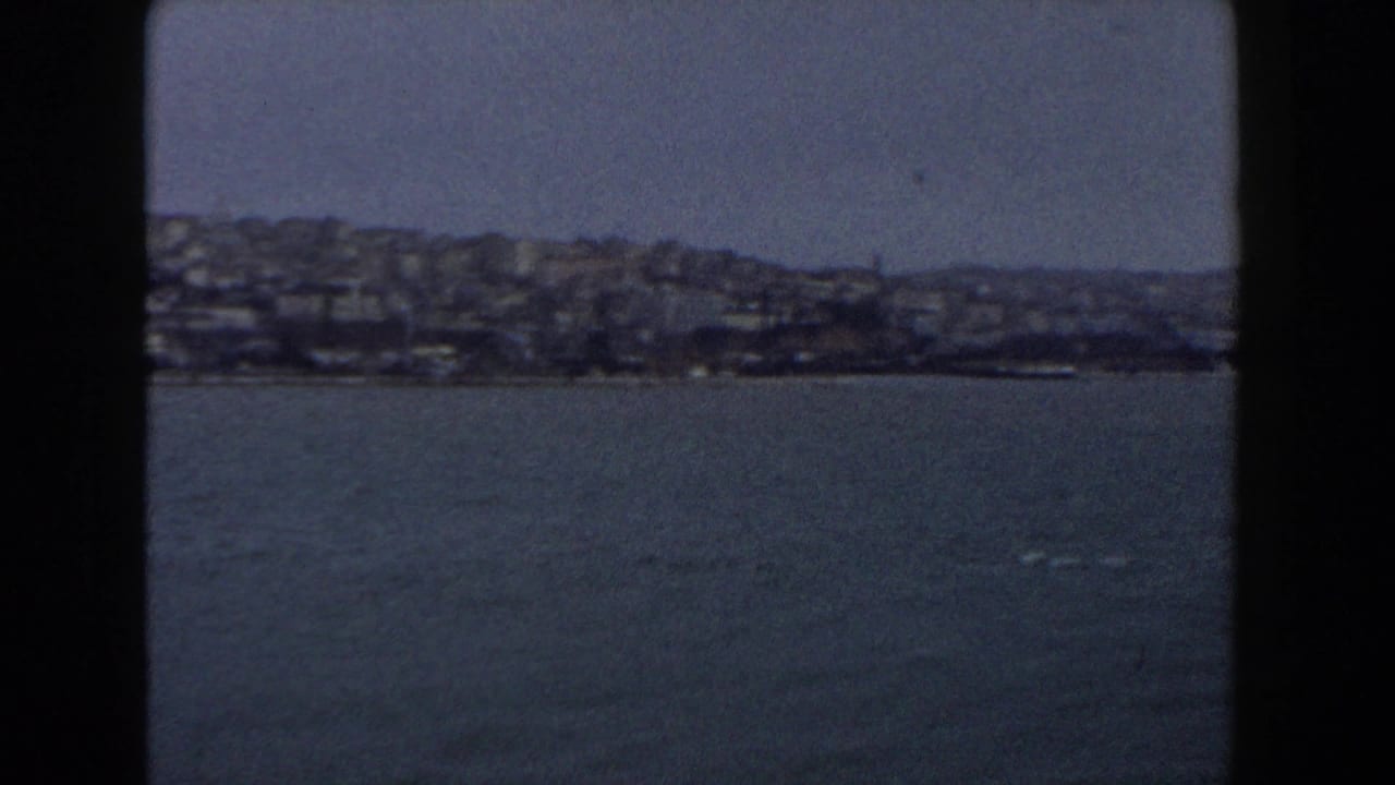 1960 Italy 8mm frame showing open water and a distant built-up hillside shoreline, with heavy gate borders and soft focus.