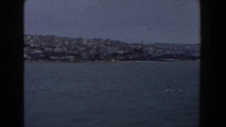 1960 Italy 8mm frame showing open water and a distant built-up hillside shoreline, with heavy gate borders and soft focus.