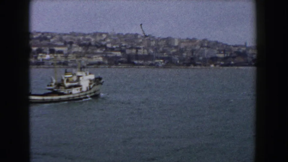 A small boat crosses open harbor water in 1960 Italy, with a dense hillside waterfront visible in the background.