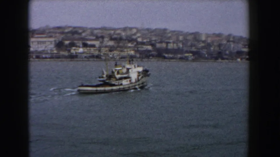 The boat continues across the water, leaving a white wake below a dense hillside shoreline in archival 8mm footage from Italy.