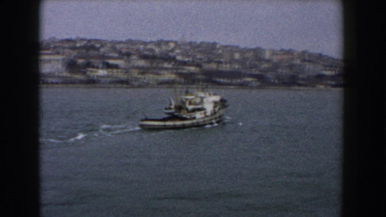 1960 Italy 8mm frame showing a small boat leaving a wake across open water below a hazy hillside city.