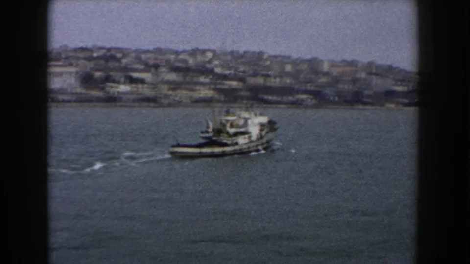 1960 Italy 8mm frame showing a small boat leaving a wake across open water below a hazy hillside city.