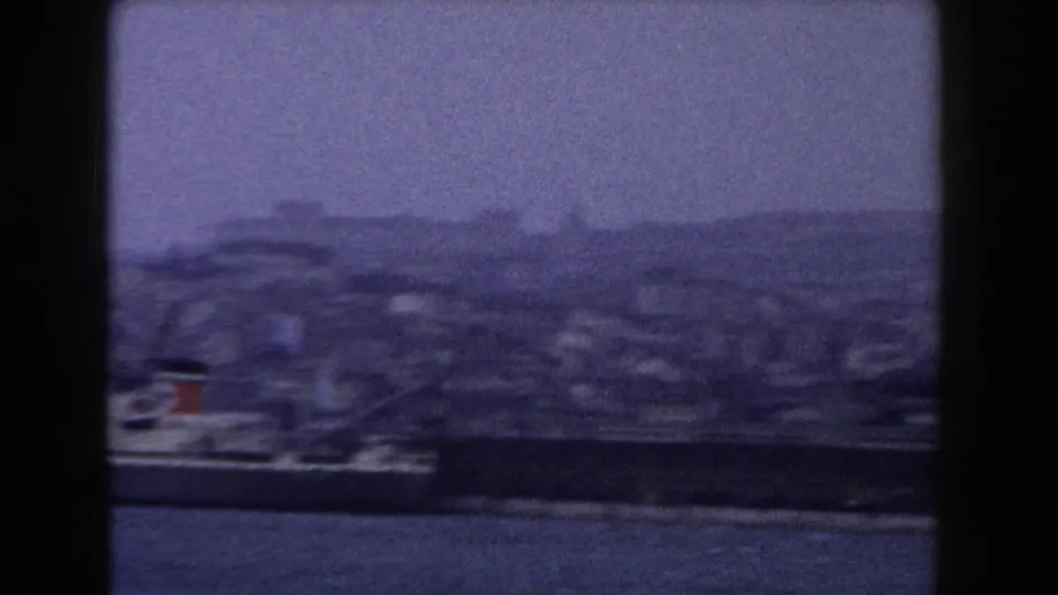 Soft 8mm view of an Italian waterfront with a docked vessel, harbor wall, and dense buildings rising behind it.