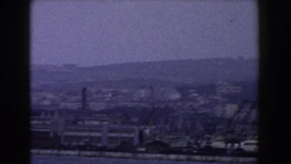 1960 Italy home-movie frame with industrial-looking waterfront structures and a hazy hillside city beyond the water.