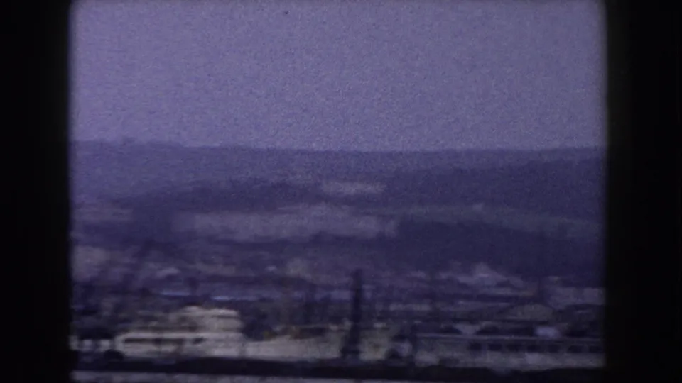 Wide 8mm coastal view in Italy with water in the foreground, low shoreline buildings, and hazy hills in the distance.