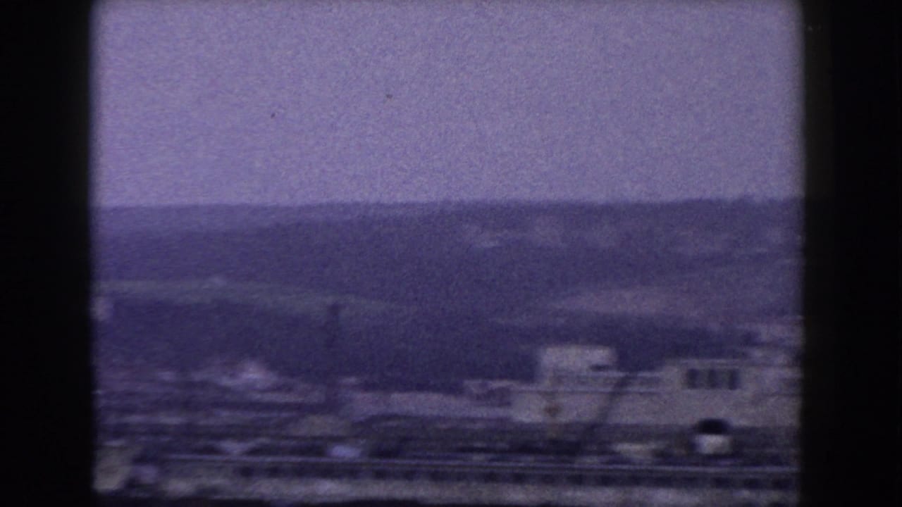 1960 8mm view of an Italian town with low buildings, cranes, hazy hills, and dark frame edges.