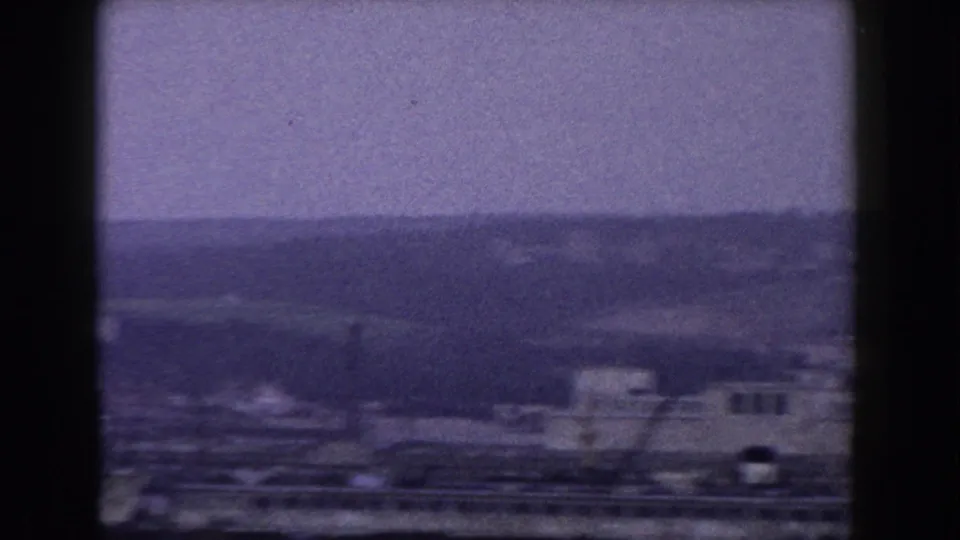 1960 8mm view of an Italian town with low buildings, cranes, hazy hills, and dark frame edges.
