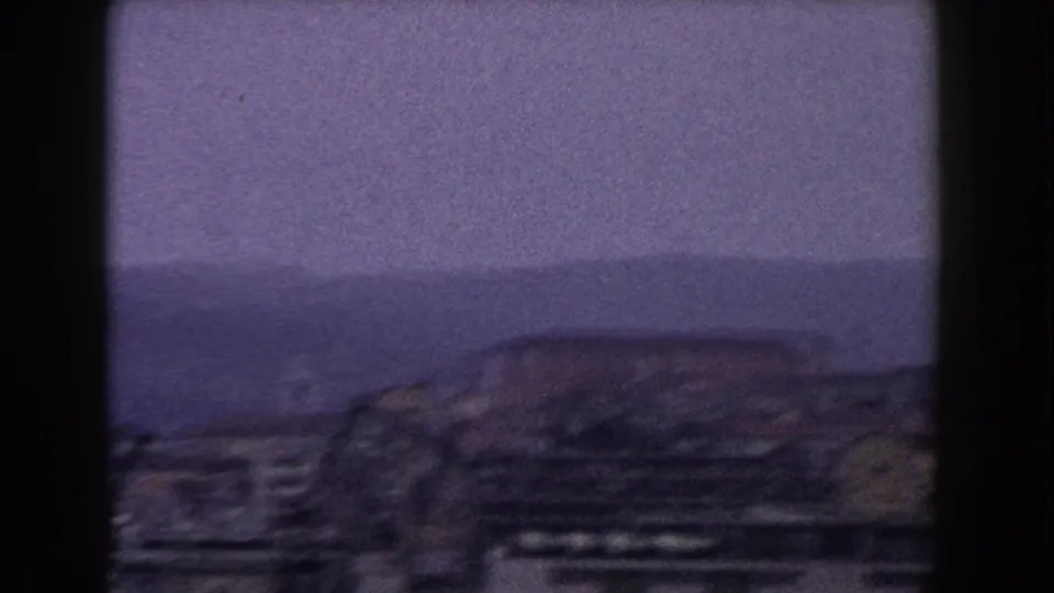 1960 Italian home-movie frame showing a large roofline, distant haze or water, and hills beyond the town.