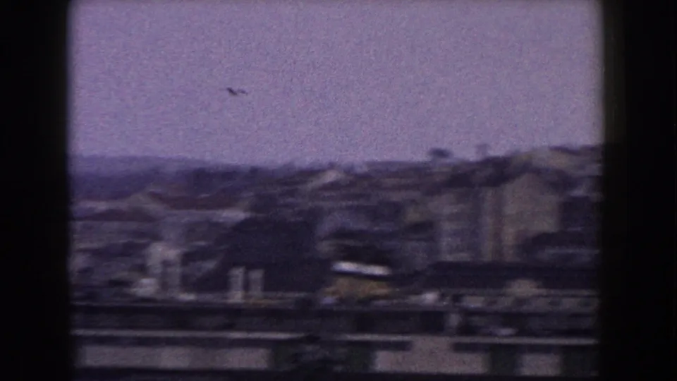 1960 8mm Italian town view with rooflines, hillside buildings, cranes, and a bird in the pale sky.