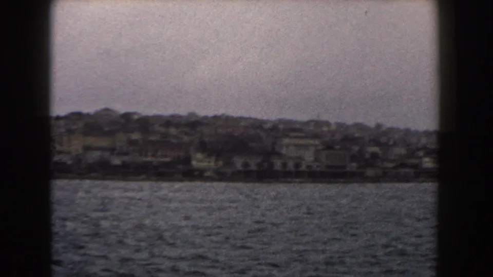 A 1960 Italian coastal or lakeside town is seen from the water, with buildings stretching along the shore.