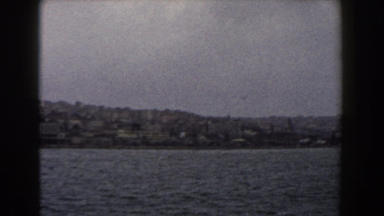 1960 Italy 8mm view of a hazy coastal city and hillside buildings seen across open water on a cloudy day.
