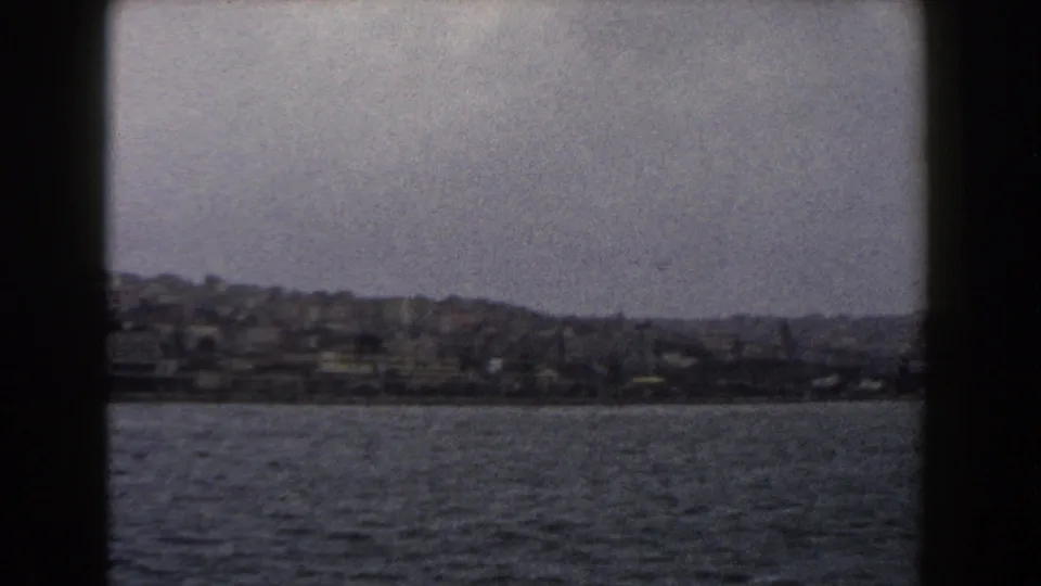 1960 Italy 8mm view of a hazy coastal city and hillside buildings seen across open water on a cloudy day.