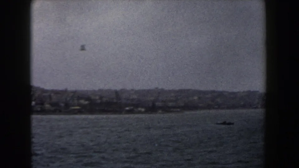 1960 Italy coastal view with dense hillside buildings, grey water, cloudy sky, and a small boat offshore.