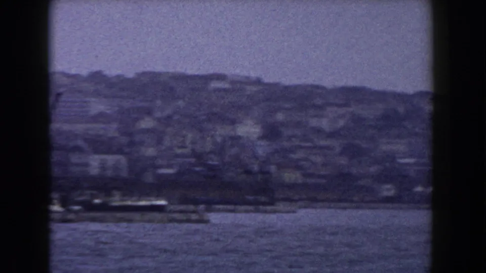Soft 8mm frame of an Italian coastal city around 1960, with waterfront structures and dense hillside buildings beyond the water.