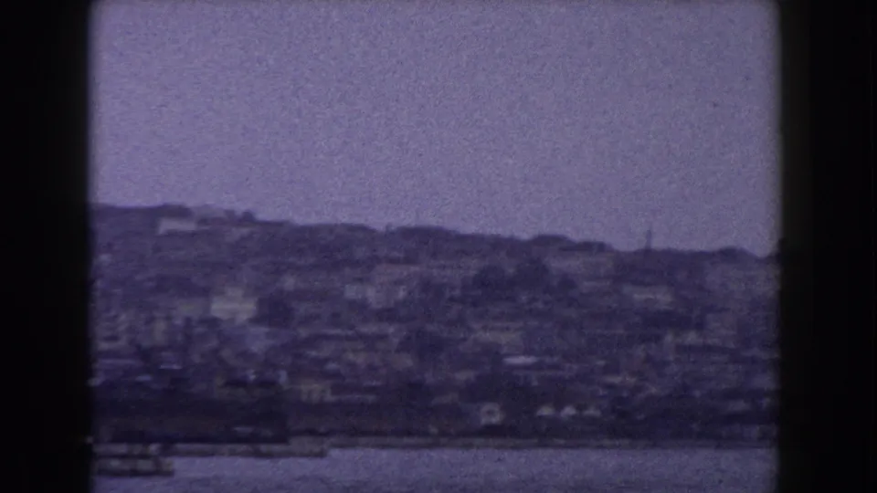 1960 Italy 8mm coastal city view showing dense hillside buildings, waterfront forms, and open water beneath a muted sky.