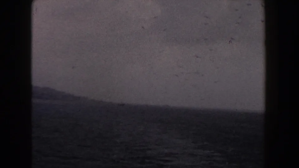1960 Italy coastal 8mm view with dark water, a distant low shoreline, cloudy sky, dust specks, and soft exposure.
