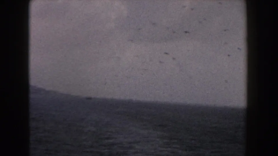 1960 Italy 8mm coastal frame with cloudy sky, dark water, a faint sloping shoreline, and visible home-movie dust marks.