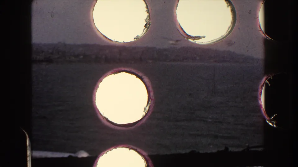 1960 Italy 8mm frame showing water and a low shoreline through dark framing, with multiple bright circular light leaks or film artifacts.