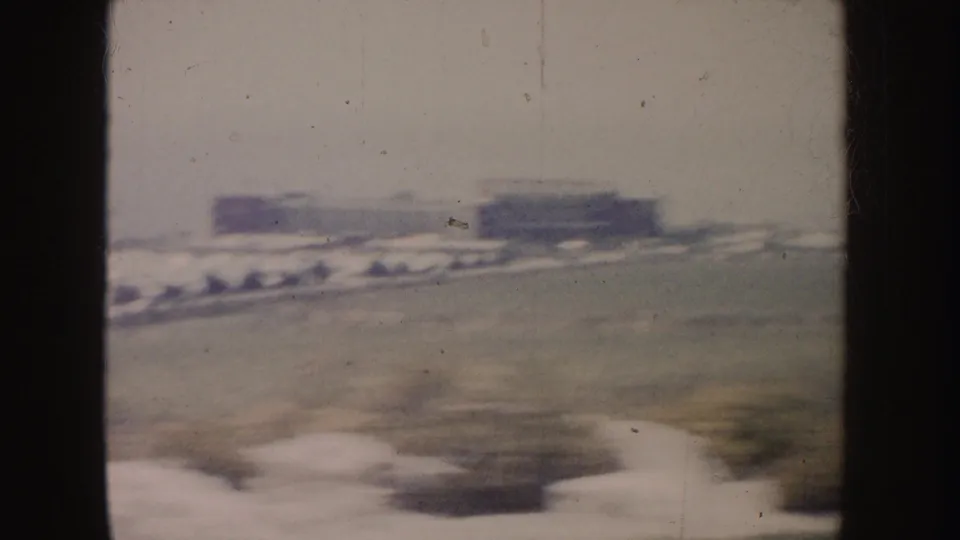 Soft 1962 California car-window view of a hazy roadside with low buildings, open ground, and a blurred passing vehicle.