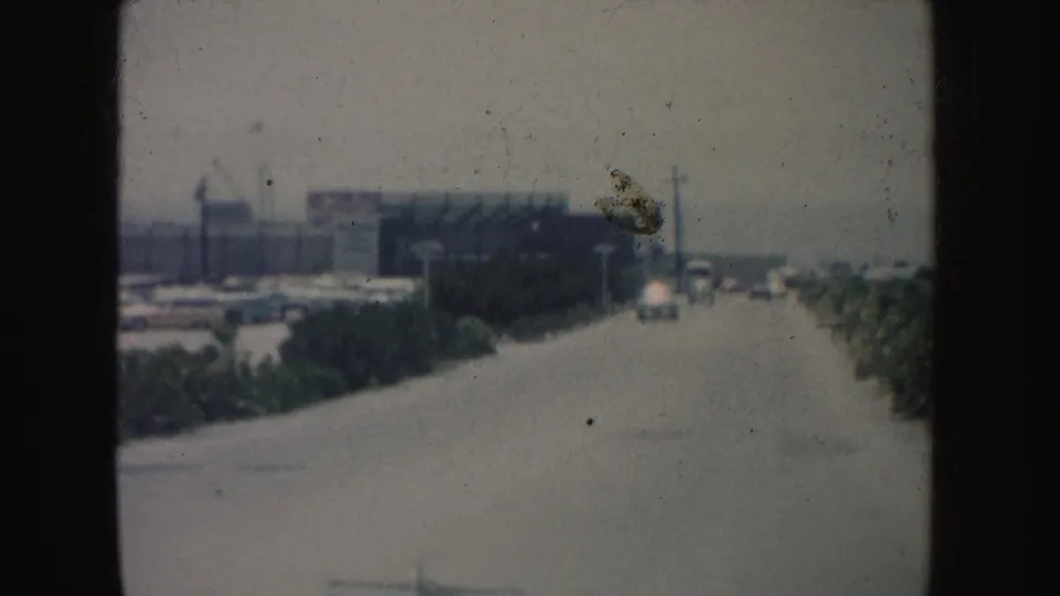 Forward-facing 1962 California road view from a car, showing roadside shrubs, utility poles, distant cars, and a low building.