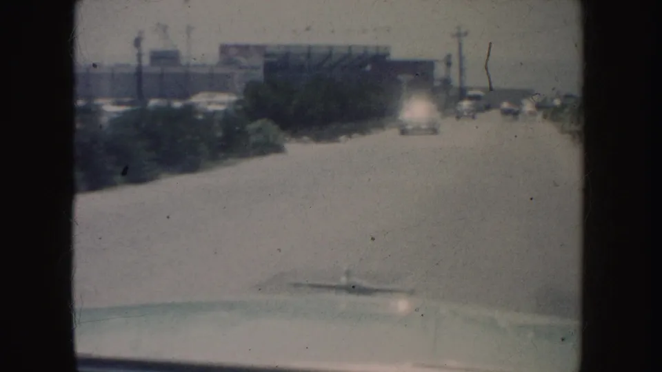 1962 California driving view with the car hood at the bottom, a pale road ahead, roadside shrubs, distant traffic, and low buildings.