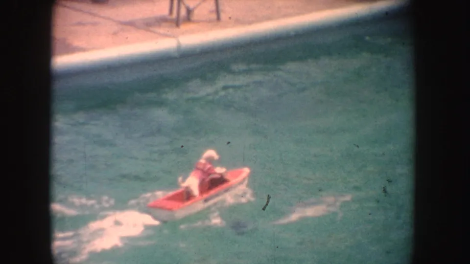 Distant person riding a small red toy boat on turquoise water in the 1962 California home-movie footage.