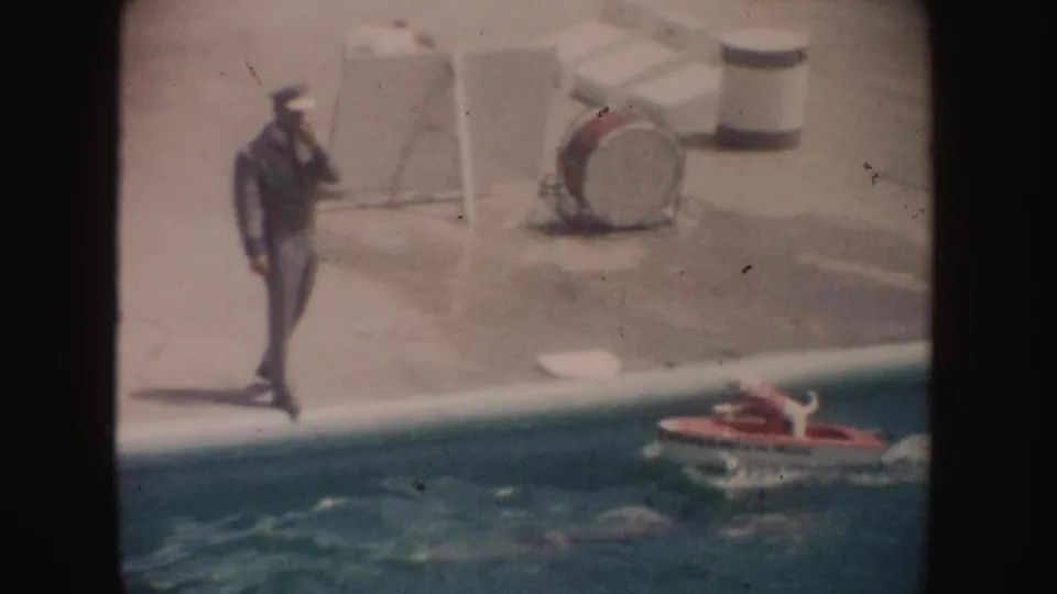 A poolside adult watches or supervises as the white dog in the red boat passes near the edge of the swimming pool.