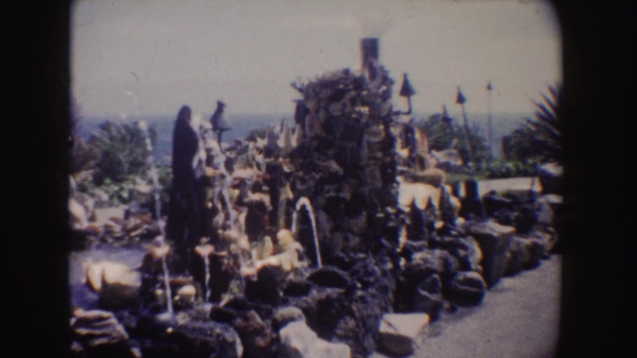 Rock fountain with water jets, small statues, lamps, ocean horizon, and paved path.