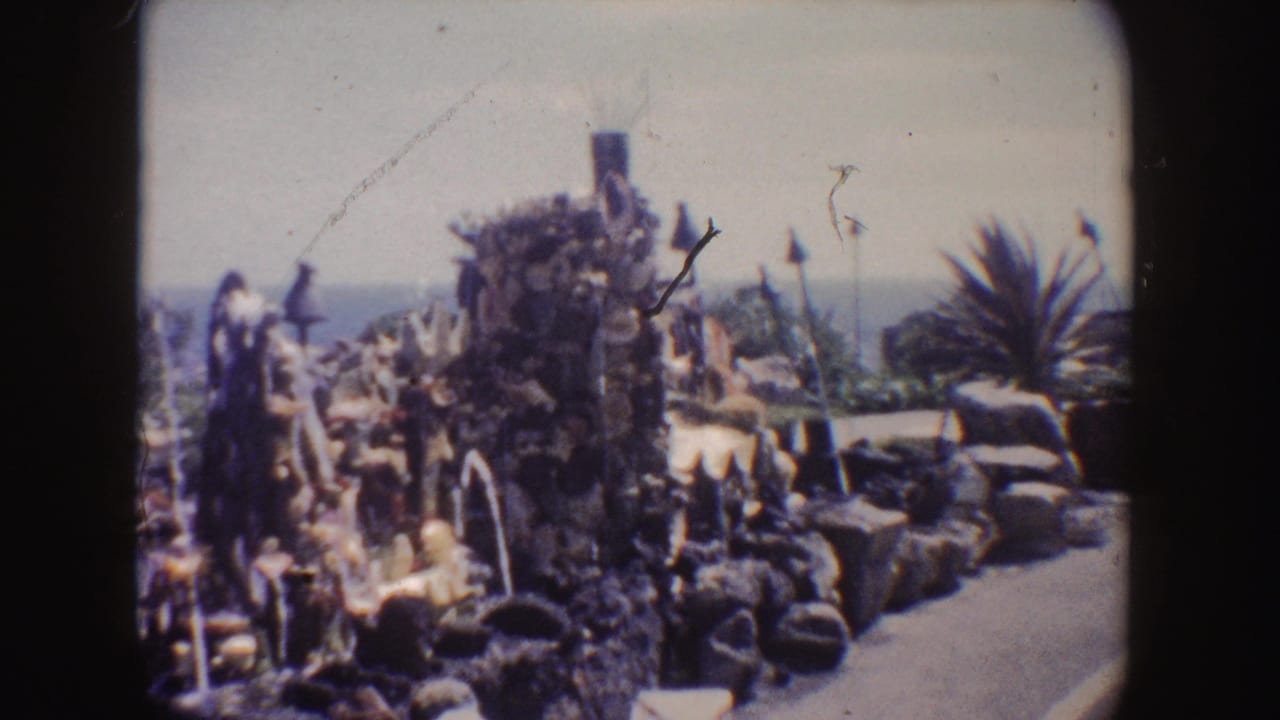 Coastal rock fountain with statues, lamp posts, palm-like planting, ocean, and walkway.