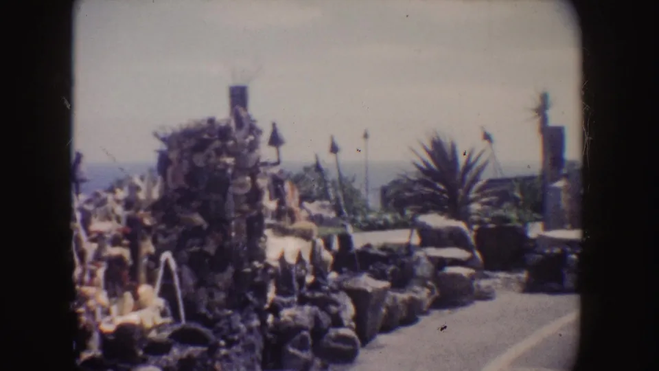 1962 California 8mm frame showing the fountain from a slightly shifted angle, including water streams, statues, palms, a column, and coastline.