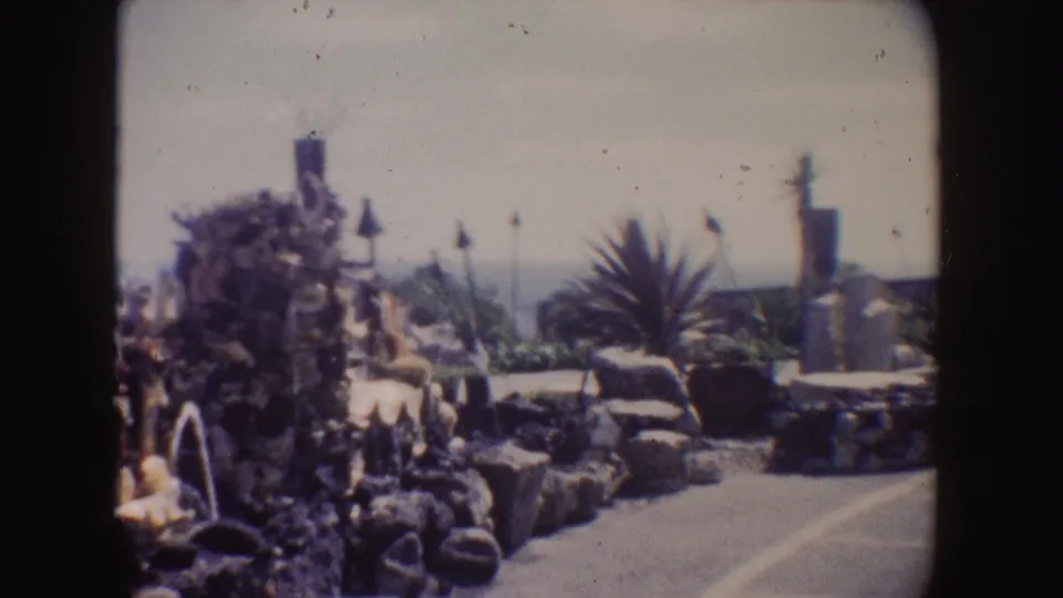 1962 California home-movie frame of the fountain display beside a walkway, with small statues, palms, a column, and ocean beyond.
