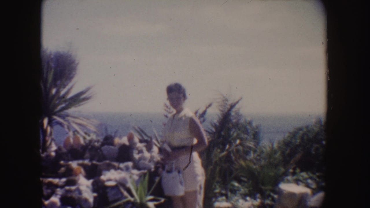 Woman in a white dress standing near garden plants with sea behind her.