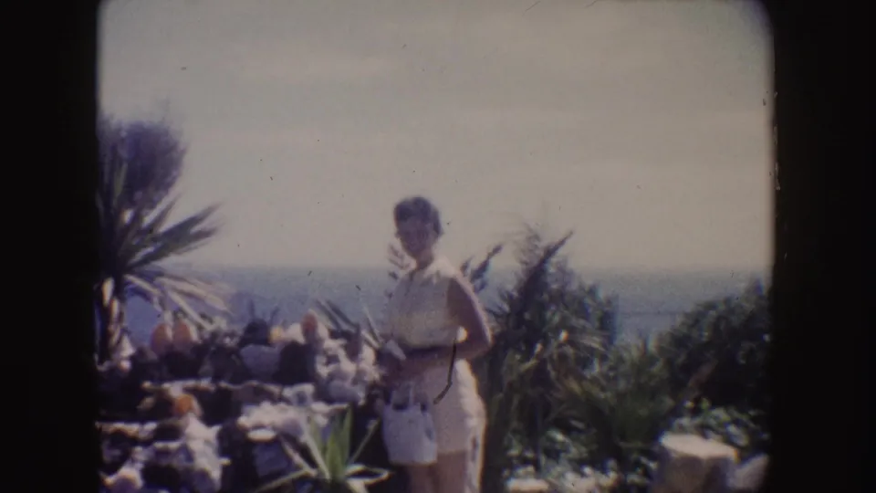 1962 California frame of a woman in a white dress posing in a coastal garden with palm-like plants and ocean in the background.