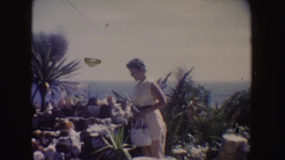 1962 California home-movie still of a woman in a white dress standing among rocks and plants above the sea.