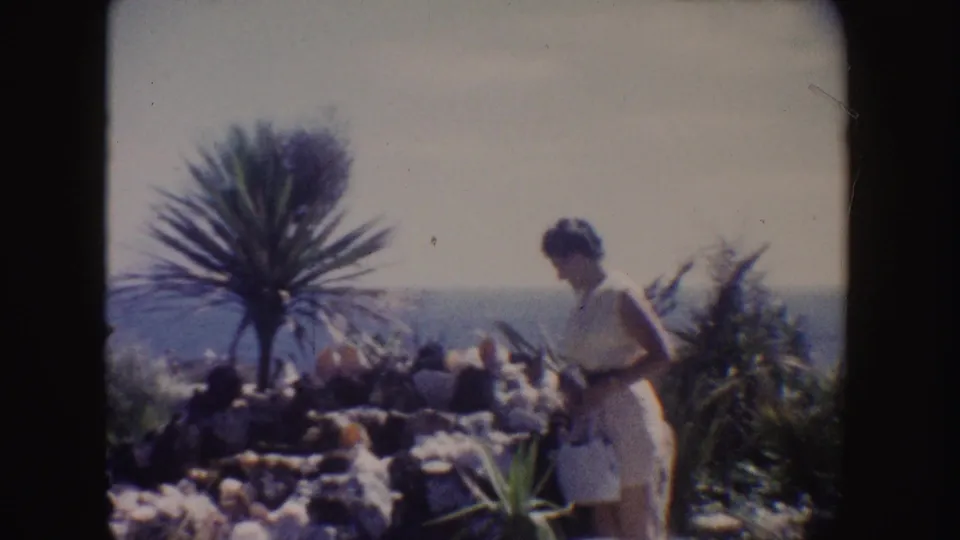 1962 California frame showing a woman in a white dress near a small palm-like plant, with rocky garden beds and ocean behind.