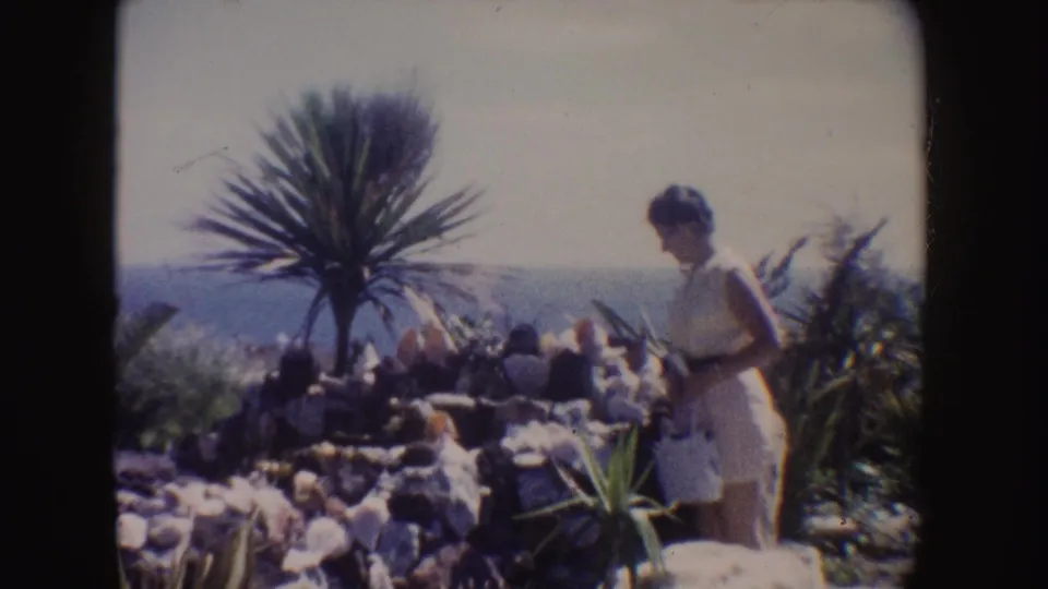 1962 California 8mm frame of a woman in a pale yellow dress on a rocky coastal overlook near yucca-like foliage and the ocean.