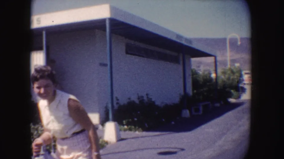 The woman moves past the camera along the paved path, with the low building, canopy posts, shrubs, and hills still visible behind her.
