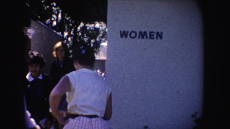 Women stand near a restroom entrance marked WOMEN in blue block letters, with trees and a building wall visible in the 1962 California frame.