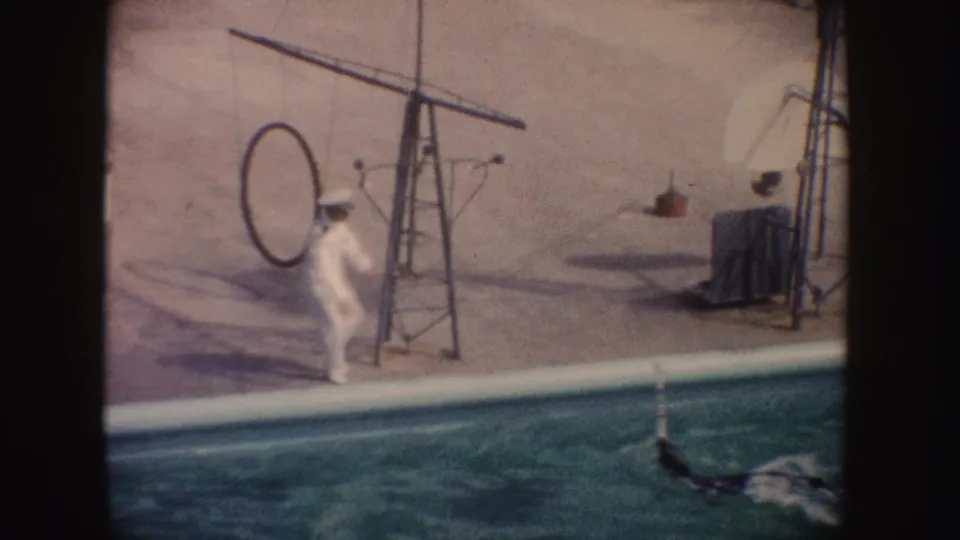 1962 California home-movie frame showing a white-uniformed man beside a marine park pool with a dolphin in the water.